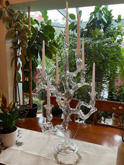 Tree of Life Candelabra In Home with white candles on a wooden table with a white cloth and many green plants in the background. Daylight comes in the back illuminating the entire scene.