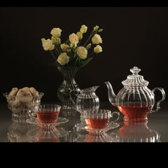 The William Teapot by Gabriela Seres, teacups on saucers, creamer, bowl and vase with yellow flowers on a reflective grey surface and black backround.