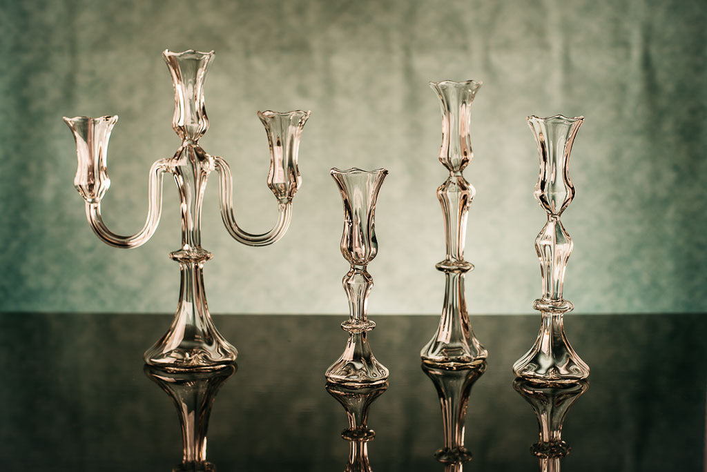 Set of four glass candelabras on a reflective surface with a greenish-brown background. Gabriela Seres designed and handmade